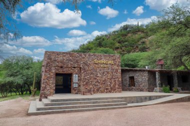 Shincal arkeoloji sahasındaki müze binası. Erişim basamakları, tabelaları ve doğal çevresiyle birlikte bitki örtüsü ve kısmen bulutlu gökyüzü olan taş yapı..