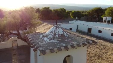 Drone, Fiambala, Catamarca 'daki San Pedro Kulesi' nin tepesindeki haçtan geri çekiliyor. Binanın havadan görünüşü, tarihi mezarlık ve sıcak gün batımı..