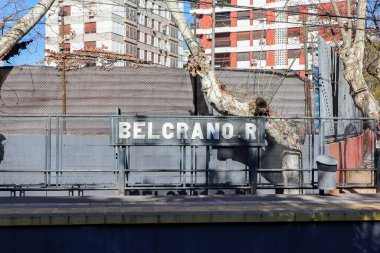 Belgrano R istasyon tabelasında, tren peronundan görülebilen tahtadan harfler var. Gri çit yapısı ve Buenos Aires 'teki Mitre demiryolu inşaatı ve ağacı ile şehir arkaplanı..