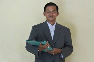 Mavi klasör tutan takım elbiseli genç bir adam. Sarı arkaplanda izole edilmiş