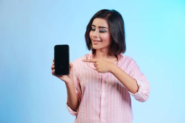 Gülümseyen sıradan bir kızın portresi mavi arkaplanda izole edilmiş boş ekran cep telefonunu işaret ediyor.