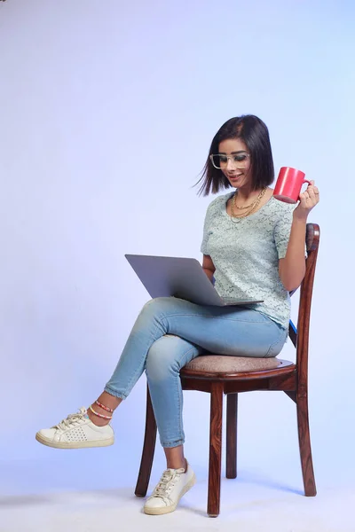 Young attractive girl with short hair, working on a laptop computer holding a red coffee mug, sitting on a wooden chair against on a studio background