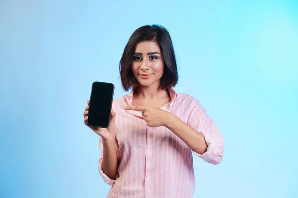一个带着微笑的随意女孩用手指指着深蓝色背景的空白屏幕手机的画像 — 图库照片
