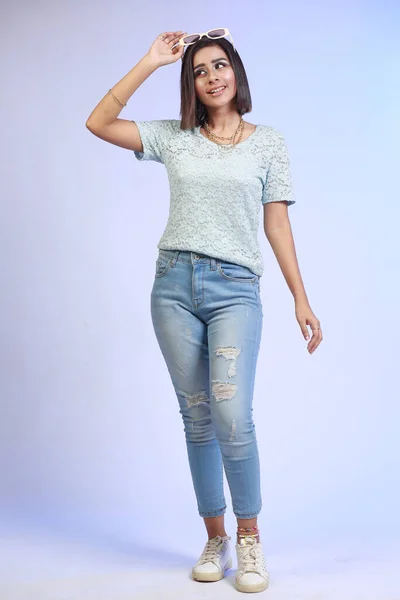 年轻迷人的巴基斯坦印度女孩 穿着牛仔裤 白鞋和时髦太阳镜 在蓝色渐变的工作室背景上摆出迷人的表情 — 图库照片