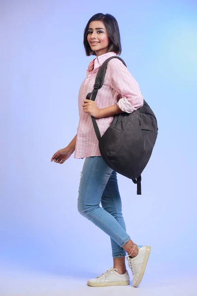 身着牛仔裤和粉色衬衫的女生背着背包在工作室背景下行走的全景照片 — 图库照片