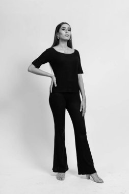 Black and white photo of a Young attractive Slim fit curvy girl posing casually, wearing black looking away from camera keeping one hand on waist. Isolated on a white studio background
