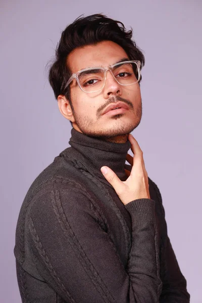 a man in a turtle neck knitted sweater and specs, touches his neck on a studio background with a serious face portrait close-up of a male model