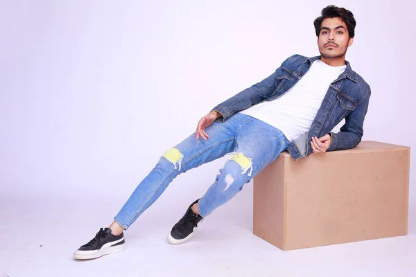 Young Asian Pakistani fashion model in a denim jacket and pants, sitting on a wooden box and posing high fashion against studio background