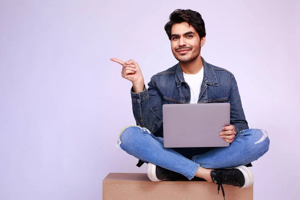 Pakistani man sitting on a box with his laptop pointing finger to the side at the copy space