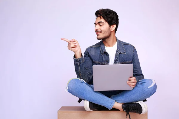 Pakistani man sitting on a box with his laptop pointing finger to the side at the copy space