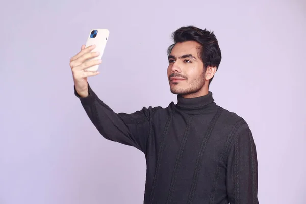 Happy successful businessman in a black knitted sweater taking selfie looking at smart phone. half body mid shot portrait isolated over light pink background.