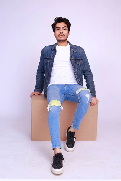 Young Asian Pakistani fashion model in a denim jacket and pants, sitting on a wooden box and posing high fashion against studio background
