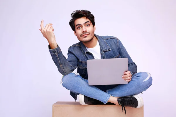Young Pakistani man in a denim jacket and pants, sitting on a box with laptop and has surprised, clueless expressions, gesture shows man questioning