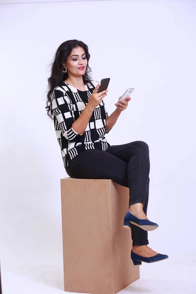 Happy stylish young brown woman using two smart phones, wearing black and white shirt with black pants sitting on a box stool