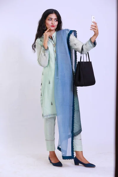 Pakistani woman descent, dressed in traditional attire, wearing a kamiz and shalwar. She has her hair open and is using a smartphone, as well as carrying a handbag. 