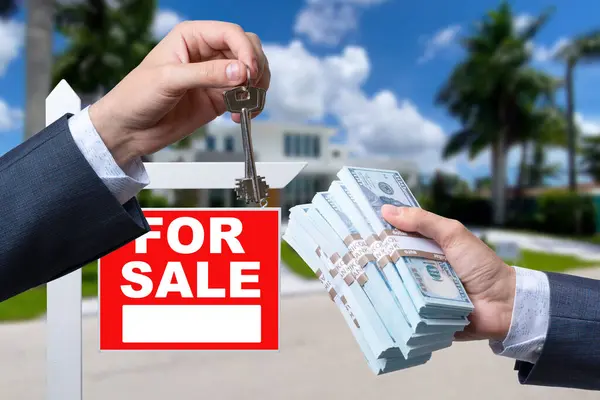 Agent Handing Over Keys as Buyer is Handing Over Cash for House with Home and For Sale Real Estate Sign Behind