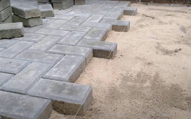 İşçiler kumlu bir yüzeye düzgün bir desen içinde gri beton kaldırımlar kuruyorlar. Bölge, istiflenmiş taşlar ve inşaat için temiz bir çalışma alanının yanında kısmen tamamlanmış kaldırımları gösteriyor..