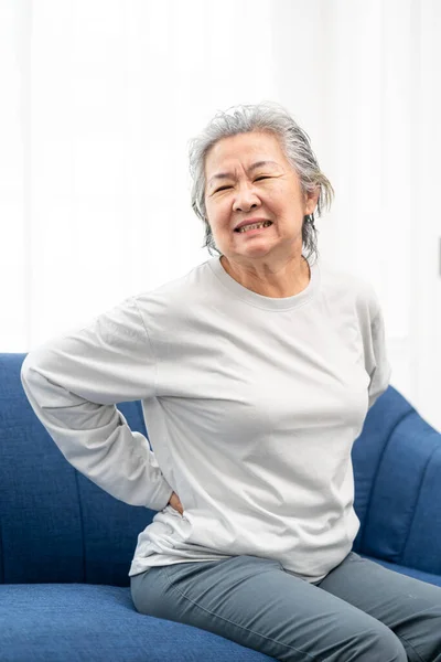 stock image senior woman sitting on a sofa at home with a backache, feeling pain and with an expression of being unwell. Health problem concept.