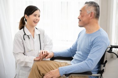 Yaşlı adam tekerlekli sandalyede otururken Asyalı kadın doktor el ele tutuşuyor. Tekerlekli sandalyedeki yaşlı adam onunla el ele tutuşurken doktorla konuşuyor..