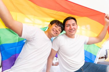 Happy Asian LGBT couple, sitting on the sofa holding and waving rainbow LGBT Pride flag together in the bedroom at home. Diversity of LGBT relationships. A gay couple concept. Loving gay couple.