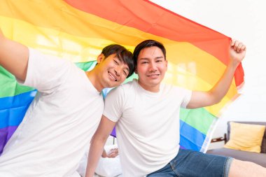 Happy Asian LGBT couple, sitting on the sofa holding and waving rainbow LGBT Pride flag together in the bedroom at home. Diversity of LGBT relationships. A gay couple concept. Loving gay couple.