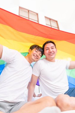 Happy Asian LGBT couple, sitting on the sofa holding and waving rainbow LGBT Pride flag together in the bedroom at home. Diversity of LGBT relationships. A gay couple concept. Loving gay couple.