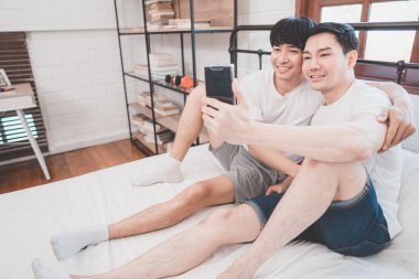 Happy Asian gay couple use mobile phone video call or selfie on bed, LGBTQ concept. Happy gay couple having fun using mobile smartphone in bed. Homosexual love and technology concept