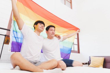 Happy Asian LGBT couple, sitting on the sofa holding and waving rainbow LGBT Pride flag together in the bedroom at home. Diversity of LGBT relationships. A gay couple concept. Loving gay couple.
