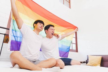 Happy Asian LGBT couple, sitting on the sofa holding and waving rainbow LGBT Pride flag together in the bedroom at home. Diversity of LGBT relationships. A gay couple concept. Loving gay couple.