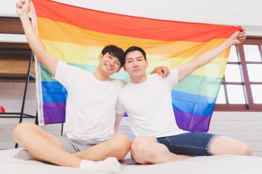 Mutlu Asyalı LGBT çifti, evdeki yatak odasında kanepede oturmuş, gökkuşağı LGBT Onur bayrağını sallıyorlar. LGBT ilişkilerinin çeşitliliği. Eşcinsel çift kavramı. Sevgi dolu eşcinsel çift.