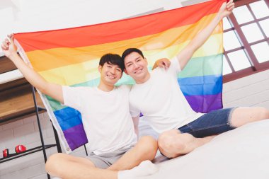 Happy Asian LGBT couple, sitting on the sofa holding and waving rainbow LGBT Pride flag together in the bedroom at home. Diversity of LGBT relationships. A gay couple concept. Loving gay couple.