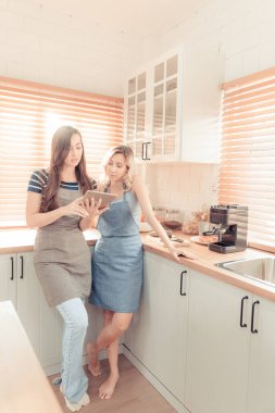 Beautiful lesbian couple cooking healthy food, looking on digital tablet at modern kitchen interior. Homosexual relations and healthy eating concept.  LGBTQ+,LGBT,LGBTQ home online kitchen. warm tone.