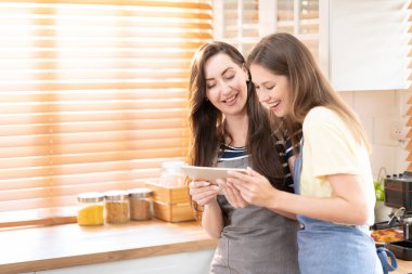 Beautiful lesbian couple cooking healthy food, looking on digital tablet at modern kitchen interior. Homosexual relations and healthy eating concept.  LGBTQ+,LGBT,LGBTQ home online kitchen.