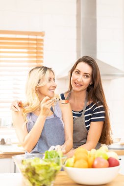 Happy same sex couple feed each other at kitchen. Cheerful LGBTQ lesbian woman feeding salad each other. A beautiful couple of lesbian ladies having a romantic moment in the kitchen.