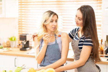 Happy same sex couple feed each other at kitchen. Cheerful LGBTQ lesbian woman feeding salad each other. A beautiful couple of lesbian ladies having a romantic moment in the kitchen.