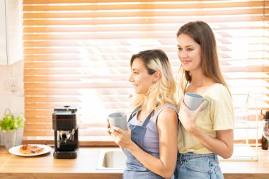 happy lesbian couple holding cups of coffee in kitchen. Couple of lesbian Girls Enjoy coffee at home taking about something. Two young adult beautiful women drink tea in modern kitchen.