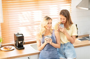 happy lesbian couple holding cups of coffee in kitchen. Couple of lesbian Girls Enjoy coffee at home taking about something. Two young adult beautiful women drink tea in modern kitchen.