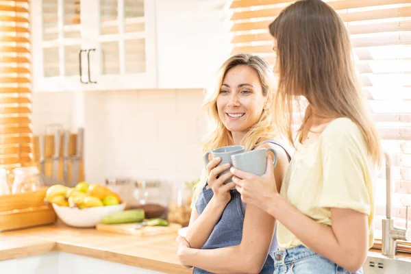 stock image happy lesbian couple holding cups of coffee in kitchen. Couple of lesbian Girls Enjoy coffee at home taking about something. Two young adult beautiful women drink tea in modern kitchen.