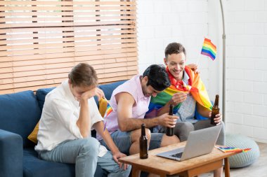 Oturma odasında oturan üç genç adam bilgisayar kullanarak gökkuşağı LGBT Onur bayrağını sallıyor, arıyor ve tutuyor. LGBTQ insan yaşam tarzı. Gurur gününü sersemletiyor..