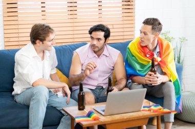 Oturma odasında oturan üç genç adam bilgisayar kullanarak gökkuşağı LGBT Onur bayrağını sallıyor, arıyor ve tutuyor. LGBTQ insan yaşam tarzı. Gurur gününü sersemletiyor..