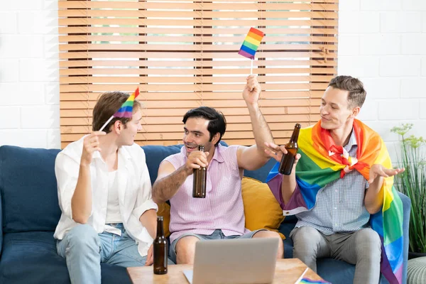 stock image Triple young man sitting in the living room while using laptop to video call and holding and waving rainbow LGBT Pride flag. LGBTQ people lifestyle. cerebrating the pride day.