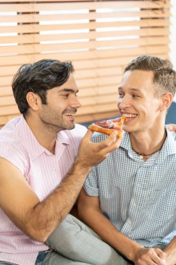 Pizza yiyen neşeli eşcinsel çift. Evdeki kanepede oturan eşcinsel çift bir dilim pizza yiyorlar. Homoseksüel ilişki kavramı.