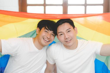 Mutlu Asyalı LGBT çifti, evdeki yatak odasında kanepede oturmuş, gökkuşağı LGBT Onur bayrağını sallıyorlar. LGBT ilişkilerinin çeşitliliği. Eşcinsel çift kavramı. Sevgi dolu eşcinsel çift.