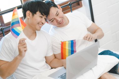 Happy engaged asian gay couple waving lgbt rainbow flag and VDO call to share happy moment at bedroom. happy married homosexual gay couple. Young Asia Gay couple feeling happy showing ring at home.