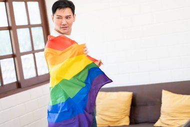 Gökkuşağı bayrağına sarılmış neşeli Asyalı eşcinsel adam evde eğleniyor. Homoseksüel Igbtiq konsepti, gökkuşağı bayrağı, kutlama geçidi. Boşluğu kopyala.