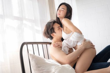 Asyalı kadın erkeğin üzerine otur ve onu kucakla. Birbirlerine bakıyorlar. Seksi tutkulu genç çift yatak odasında sevişmeden önce yatakta sarılıyor. Sevgililer Günü.