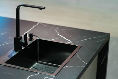 Pürüzsüz ve modern mutfak lavabosu ve musluğu karanlık bir dolabın ve mermer bir tezgahın üzerine kurulmuş. Musluk, uzun, kemerli ve siyah kaplamalı tek kollu bir tasarım..