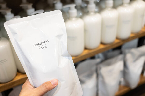 Hand Holding Shampoo Refill Pouch with Bottles on Shelf, Eco-Friendly Packaging Concept. Sustainable packaging, zero waste shopping, and eco conscious personal care.