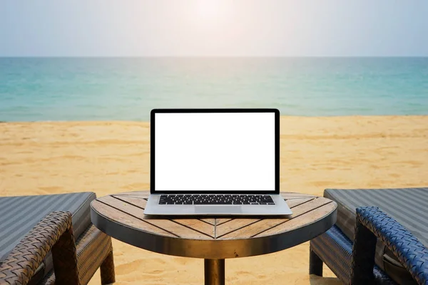 Laptop with blank screen for creative design on the table nearby sea and sand beach background with sun ray effect. Computer notebook with monitor clipping path for present landing page design. Laptop computer mock up template