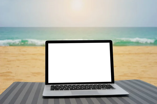 Laptop with blank screen for creative design on beach bed nearby beach and blue sea background with sunlight effect. Computer with clipping path for present landing page design mock up template